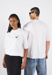 Vita kortärmade t-shirts med skorpionbroderi på framsidan, bärs med en svart kjol och blå jeans, på två stående figurer.