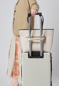 Beżowa torba na ramię w wzory z brązowymi uchwytami leży na białej walizce z żebrowanym wzorem, która ma czarną podstawę i wysuwany uchwyt.