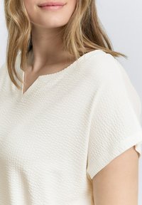 Wafeltextuur, off-white top met een V-hals en korte mouwen. De stof lijkt lichtgewicht en ademend, geschikt voor casual dragen.
