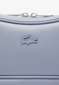 Borsa in pelle texturizzata blu chiaro con design curvo. Presenta un logo di coccodrillo in rilievo ben visibile. Chiusura con zip e dettagli in hardware argento.