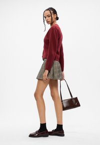 Κόκκινο πλεκτό πουλόβερ, καρό μίνι φούστα, μαύρες κάλτσες και γυαλιστερά μπορντό μοκασίνια, κρατώντας μια καφέ τσάντα χειρός με δομημένο σχέδιο.