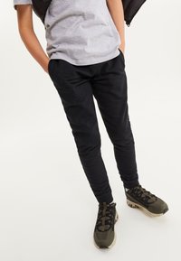 Pantalon de jogging noir à coupe slim, fabriqué en tissu doux. Associé à des chaussures de sport vertes et noires texturées avec des semelles distinctives.