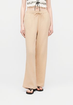 Woman wearing beige wide-leg pants with drawstring waist, a cropped ruffled top, and black thong sandals, standing against a plain background.