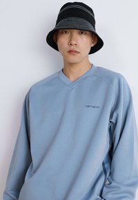 Light blue sweatshirt with a V-neck and ribbed cuffs, featuring a small black 'Carhartt' logo. Paired with a black striped bucket hat.