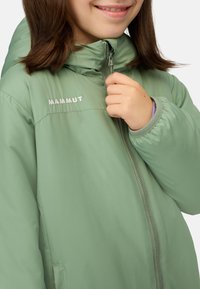 Giacca puffer verde chiaro con chiusura lampo frontale, colletto alto e logo ricamato. Presenta una texture liscia e lucida e polsini elasticizzati.