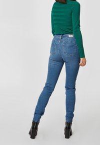 Femme portant un jean bleu ajusté, un haut vert à manches longues et des bottines noires à talons, debout de dos face au spectateur.