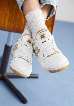Osoba nosząca białe i beżowe sneakersy New Balance 550 z białymi prążkowanymi skarpetkami, siedząca na drewnianym krześle na jasnoniebieskiej podłodze.