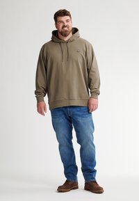 Felpa con cappuccio verde oliva, con lacci, vestibilità rilassata, abbinata a jeans blu e stivali marroni, in piedi contro uno sfondo neutro.