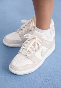 Tenisky Nike v krémových a béžových tónech, s texturovanými překryvy, perforovanou špičkou a plochými tkaničkami, se stříbrným akcentem na jazyku.