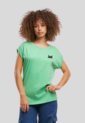 Femme aux cheveux bouclés portant un t-shirt vert avec un petit motif de chat noir sur la poitrine et un pantalon bleu, debout avec une main sur la hanche.