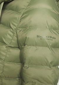 Puffjakke i lys olivengrøn, lavet af skinnende materiale med quiltede sektioner. Har logoet "Marc O'Polo" i mørkegrøn på stoffet.