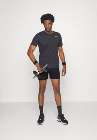 Schwarzes T-Shirt mit kurzen Ärmeln und weißem Logo, schwarze Shorts und schwarze Sportschuhe. Hält eine Kurzhantel, trägt Handgelenkbänder.
