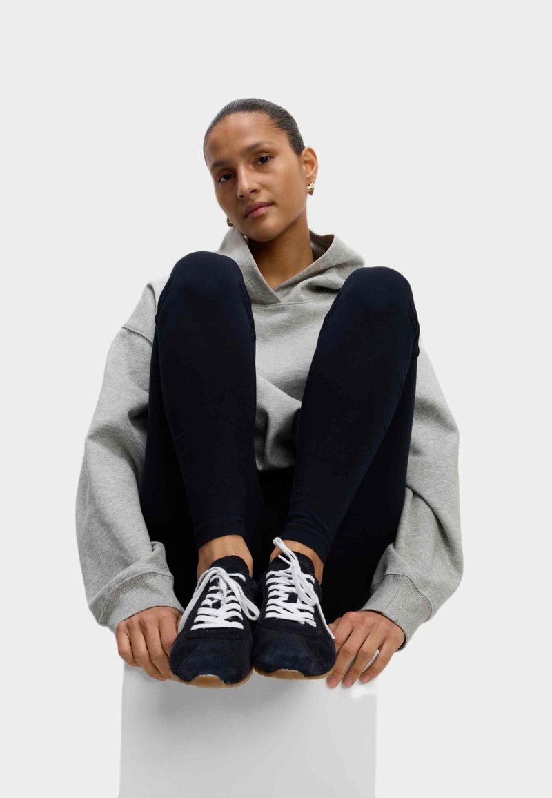 Grijze hoodie, zwarte leggings en zwarte sneakers met witte veters. Sneakers hebben een gumrubberen zool en een zachte suède structuur.