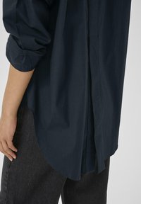 Mørkeblå skjorte med lange ærmer, rundet kant og en rygfold. Båret over sorte jeans, der fremviser et glat, tekstureret stof.