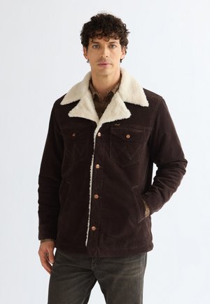 Homme aux cheveux bouclés portant une veste marron foncé avec un col en peau de mouton blanc et une chemise à carreaux, debout devant un fond blanc uni.