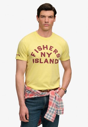 Homme portant un t-shirt jaune avec le texte "FISHERS NY ISLAND", un jean bleu, une chemise à carreaux nouée autour de la taille, et une montre-bracelet.