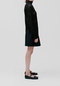 Schwarzes Langarm-Top mit Stehkragen und transparentem Material, kombiniert mit einem schwarzen A-Linien-Rock und schwarzen flachen Schuhen mit Schnallendetail.