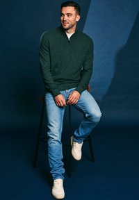 Donkergroene trui met rits gedragen door een persoon zittend op een kruk, gecombineerd met lichtblauwe spijkerbroek en witte sneakers tegen een blauwe achtergrond.