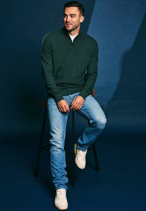 Donkergroene trui met rits gedragen door een persoon zittend op een kruk, gecombineerd met lichtblauwe spijkerbroek en witte sneakers tegen een blauwe achtergrond.