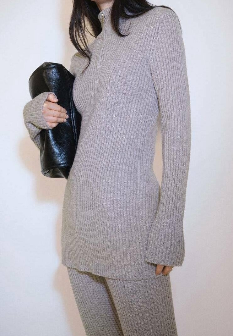 Tunique en tricot grise côtelée avec un col haut et des manches longues, associée à un sac en cuir noir. Texture lisse, silhouette ajustée.