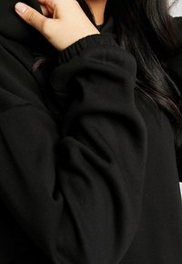 Sudadera negra con textura suave, mangas fruncidas y un ajuste relajado, mostrando pliegues y detalles de tela lisa.