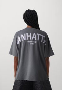 Szara bawełniana koszulka z krótkim rękawem, z napisem "MANHATTAN" oraz "New York City" w wyraźnym białym druku na plecach.