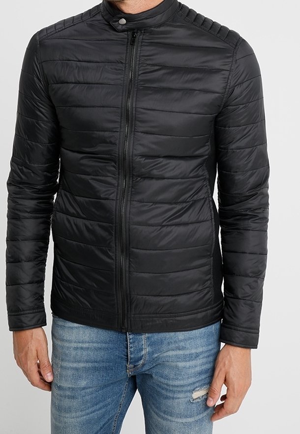 Zwarte pufferjack met een rits, verticale quiltpatroon, staande kraag en geribbelde accenten op de schouders, gecombineerd met blauwe jeans.