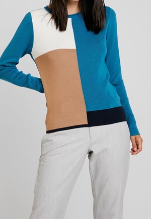 Kvinde iført en farveblok-sweater i grønblå, beige, hvid og marineblå kombineret med lysegrå bukser, stående med den ene hånd på hoften.