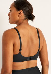 Musta rintaliivi, jossa on säädettävät hihnat, pehmeä kankaan rakenne ja hakaset takana. Yksinkertainen muotoilu ilman näkyviä kuvioita.