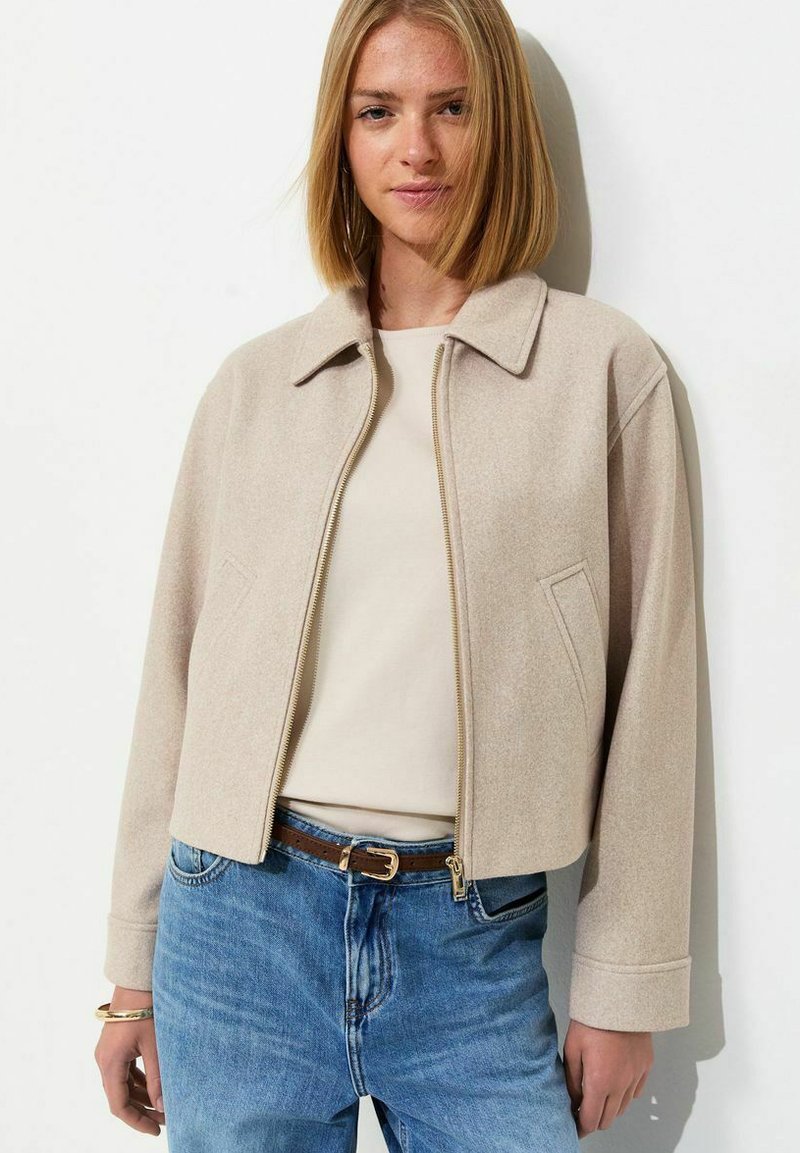 Young woman with shoulder-length blonde hair wearing a beige zip-up jacket, cream top, blue jeans, and a brown belt, standing against a white wall.