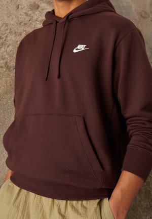 Osoba ubrana w ciemnobrązową bluzę z kapturem Nike z logo na piersi i rękami w przedniej kieszeni, w połączeniu z jasnobeżowymi spodniami na tle teksturowanej ściany.