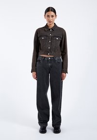 Ung kvinde med mørkt hår i en lav knold bærer en sort cropped denimjakke, løse sorte jeans og sorte sko, og står foran en hvid baggrund.