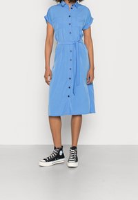 Hellblaues Hemdkleid mit kurzen Ärmeln, zwei Brusttaschen, einem Taillengürtel und Midi-Länge. Kombiniert mit schwarzen High-Top-Sneakers.