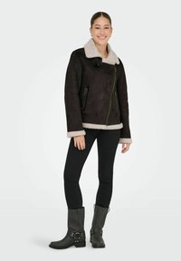 Bruine faux suede jas met een roomkleurige schapenvacht kraag en manchetten, voorzien van twee ritszakken en een diagonale rits aan de voorkant. Zwarte skinny jeans en grijze laarzen.
