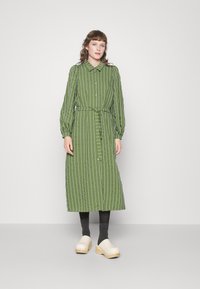 Vestido camiseiro riscado a verde, com mangas longas, cintura ajustada e botões. Usado com tamancos claros e meias escuras, em pé sobre um fundo neutro.