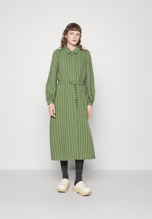 Vestido camiseiro riscado a verde, com mangas longas, cintura ajustada e botões. Usado com tamancos claros e meias escuras, em pé sobre um fundo neutro.