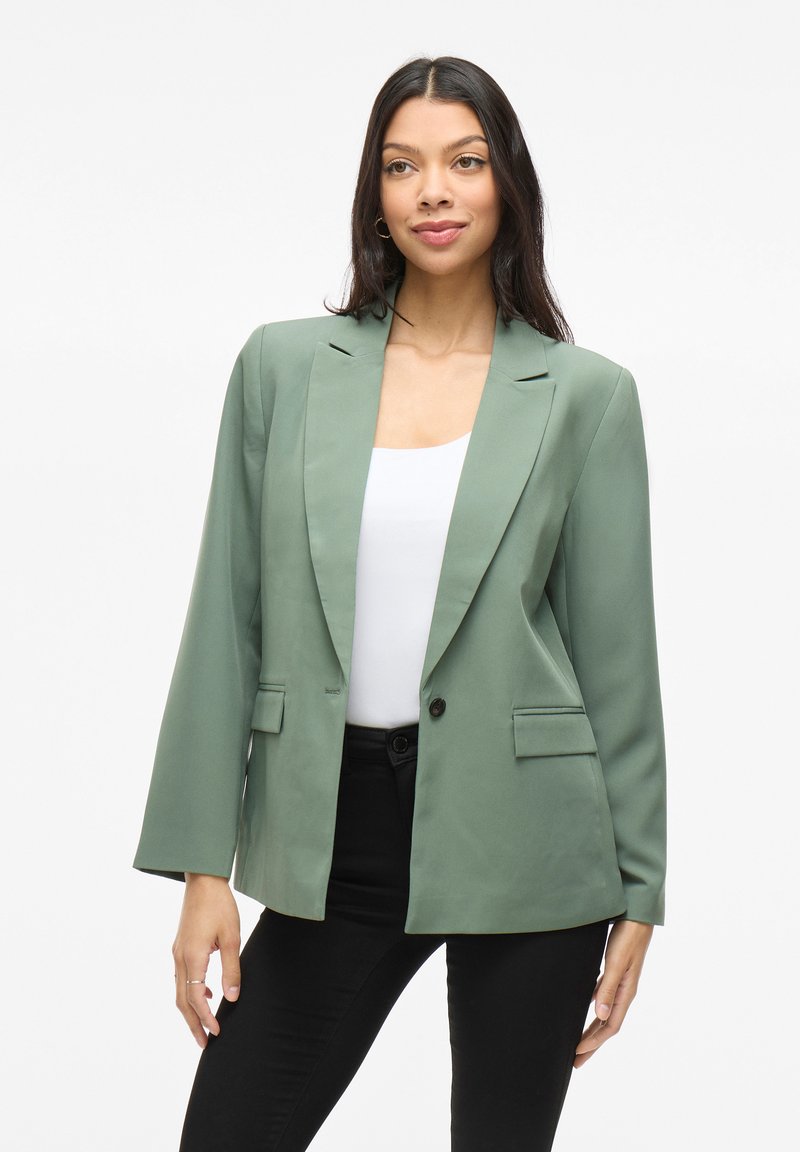 Blazer vert avec revers en cravate et fermeture à un bouton. Dispose de deux poches avant et de manches longues. Tissu lisse et coupe structurée.
