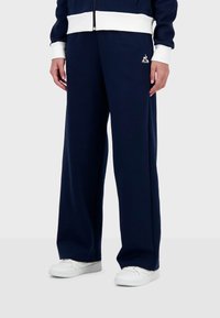 Pantalones de chándal de pierna ancha en azul marino de tejido suave, con una cinturilla elástica blanca. Detalle de pequeño logo cerca de la cadera izquierda. Llevados con zapatillas blancas.