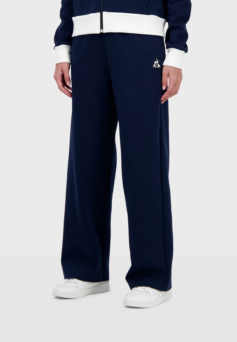 Pantalones de chándal de pierna ancha en azul marino de tejido suave, con una cinturilla elástica blanca. Detalle de pequeño logo cerca de la cadera izquierda. Llevados con zapatillas blancas.