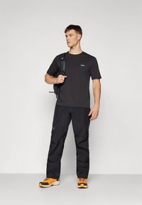 Juoda trumpomis rankovėmis palaidinė su mažu kišenės logotipu, derinama su juodomis kelnėmis ir sportiniais bateliais su oranžiniais akcentais. Kuprinė ant peties.