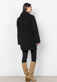Abrigo negro texturizado con cuello, de corte suelto y de longitud media. Combinado con pantalones negros ajustados y botas de rodilla beige.