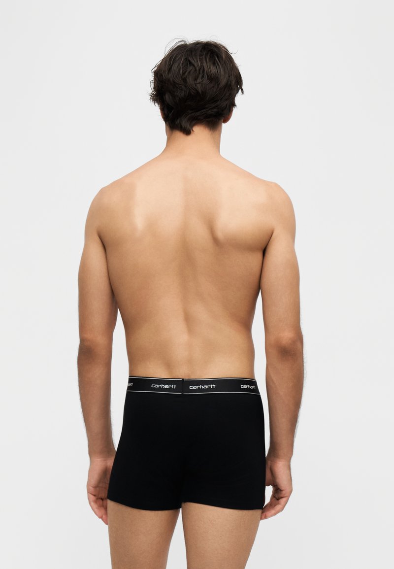 Mann mit dunklen Haaren, von hinten gesehen, trägt schwarze Carhartt-Boxershorts vor einem schlichten hellen Hintergrund.