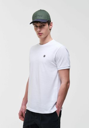 Mladi moški, ki nosi belo majico s majhnim logotipom na prsih, črne hlače in zeleno-modro baseball kapo, pozira na enobarvni podlagi.