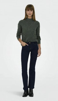 Groene gebreide trui met hoge hals, donkerblauwe wijde jeans en zwarte enkellaarsjes. De outfit heeft een aangesloten silhouet en een textuurrijke stof.