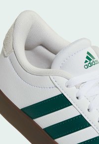 Baskets blanches Adidas avec des bandes vertes, un col rembourré, des lacets blancs et une semelle marron, vues en gros plan en angle.