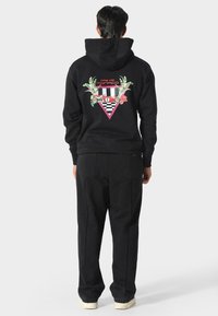 Felpa nera con un design floreale e geometrico sulla schiena, con accenti rosa e una texture morbida. Abbinata a pantaloni della tuta neri.