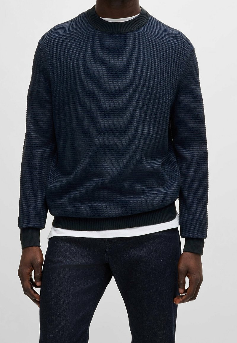 Homme portant un pull texturé bleu foncé sur une chemise blanche, assorti à un jean foncé, debout devant un fond clair uni.