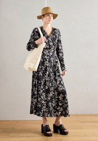 Vestido maxi de estampado floral negro con mangas largas, cintura ajustada y falda fluida. Combinado con sandalias negras de tacón bloque y un bolso tote beige.