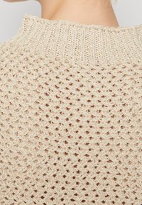Beige Strickpullover mit einem strukturierten, offenen Muster und einem hohen Kragen. Das Material wirkt leicht und hat einen leicht glänzenden Finish.