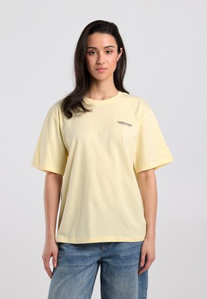 Vrouw met lang donker haar, een lichtgeel oversized T-shirt en blauwe jeans, staand tegen een effen lichtgrijze achtergrond.